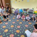 Dzieci wraz z rodzicami słuchają wiersza czytanego przez nauczycielkę